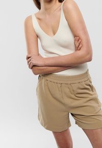 Person wearing a sleeveless, ribbed white top and beige elastic-waist shorts with hands crossed at the waist against a plain background.