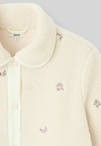 Veste chemise en polaire couleur crème avec une texture douce, col arrondi, fermeture à boutons et accent de broderies florales violettes.