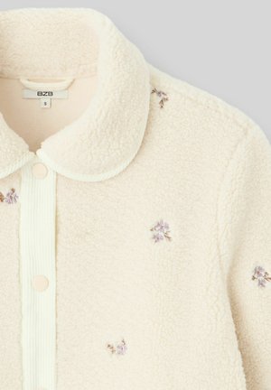 Veste chemise en polaire couleur crème avec une texture douce, col arrondi, fermeture à boutons et accent de broderies florales violettes.