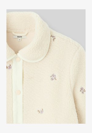 Veste chemise en polaire couleur crème avec une texture douce, col arrondi, fermeture à boutons et accent de broderies florales violettes.