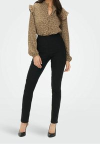 Blouse à manches longues brun à motifs avec volants aux épaules, associée à un pantalon slim noir taille haute et des chaussures à talons hauts noires.