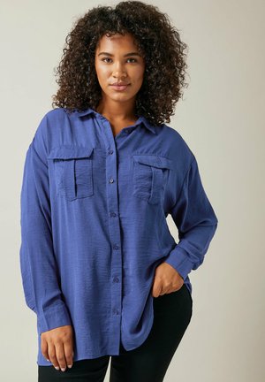 Camisa azul de botones con dos bolsillos en el pecho, mangas largas y un corte relajado. Tejido texturizado y dobladillo curvado. Llevada con pantalones negros.