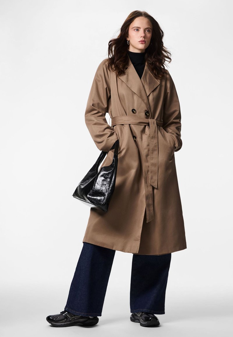 Beige trenchcoat med dobbeltradet knappelukking, belte i midjen, store slag og sidelommer. Kombinert med mørke vide jeans og en svart håndveske.