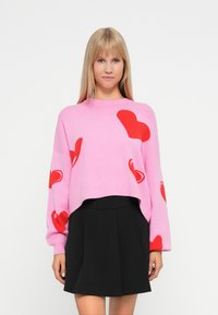 Pink cropped sweater with red heart patterns, knitted texture; paired with a black pleated skirt. Long sleeves and round neckline.