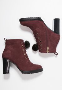 Botines burdeos de ante con ojales dorados, cordones delanteros, pompones de peluche sintético, cremallera lateral y un tacón bloque negro brillante.