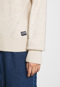 Pull beige clair avec une finition texturée et des poignets côtelés. Il présente une étiquette noire à l'ourlet, associé à un pantalon en denim bleu foncé.