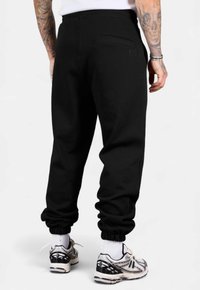 Schwarze Jogginghosen mit elastischen Bündchen und einem elastischen Bund. Verfügt über eine Gesäßtasche, eine glatte Stofftextur und eine lockere Passform. Kombiniert mit Sneakers.