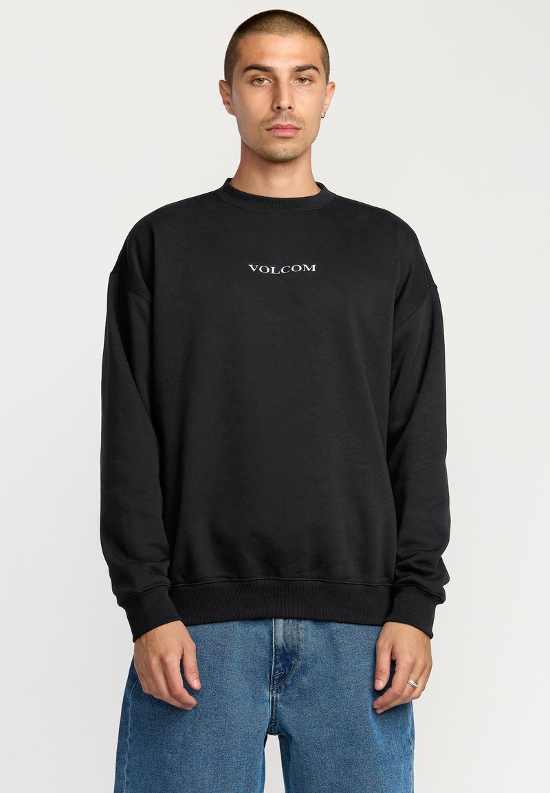 Jeune homme aux cheveux courts portant un sweat-shirt noir Volcom et un jean bleu, debout devant un fond blanc uni.