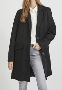 Cappotto di lana nero con colletto a rever, chiusura con un bottone, due tasche frontali e una vestibilità rilassata, indossato sopra una camicia bianca e jeans grigi.