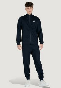 Tuta in nylon blu navy realizzata in tessuto morbido, con giacca con zip e colletto alto e pantaloni. Include il logo EAZ sul petto.