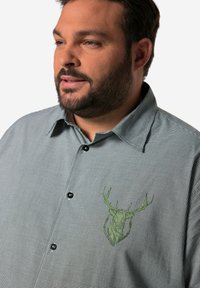Camisa de cuadros en blanco y negro con un gráfico de una cabeza de ciervo verde en el lado izquierdo del pecho. Presenta un collar con puntas y botones negros.