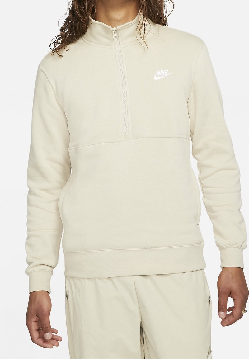 Světlé béžové mikina Nike s polovičním zipem u límce, měkkou texturou a bílým logem na hrudi; má žebrované manžety a volný střih.