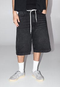Pantaloni shorts in denim nero con cordoncino bianco, taglio dritto, orlo sfrangiato, abbinati a scarpe da ginnastica in suede grigio chiaro e calze bianche a crew.