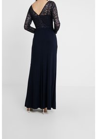 Dlouhé námořnické večerní šaty s dlouhými krajkovými rukávy, designem do V na zadní straně, přiléhavým korzetem a splývavou sukňou, se složitými krajkovými detaily v horní části.