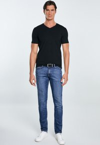 Mees seisab näoga ettepoole, seljas must lühikeste varrukatega V-kaelusega särk, sinised teksapüksid musta vööga ja valged tossud, taust on ühtlane.