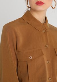 Donna che indossa una camicia marrone con bottoni e tasche a patta, con orecchini a cerchio dorati, mostrata dalle spalle alle labbra.
