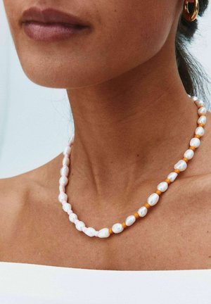 Lähikuva henkilöstä, joka käyttää helmikaulakorua, jossa on oransseja helmiä, kultaisia rengaskorvakoruja ja valkoista olkapäätöntä toppia.