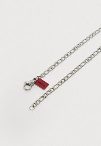 Ασημένια μεταλλική αλυσίδα με οβάλ συνδέσμους και κούμπωμα καβουριού, με κόκκινη ετικέτα που φέρει την εγχάραξη "HUGO". Ο σχεδιασμός είναι απλός και λειτουργικός.