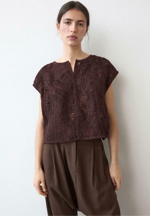 Młoda kobieta o ciemnych włosach ma na sobie teksturowany, brązowy, krótkim rękawem top wykonany na szydełku oraz luźne, brązowe spodnie, stojąc przy gładkiej białej ścianie.