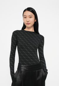 Giovane donna che indossa un top lungo attillato nero con un sottile motivo a diamante e pantaloni satinati neri a vita alta, con le mani nelle tasche.