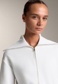 Giacca bianca con zip e colletto alto, texture liscia e dettagli in zip argentata, ambientata su uno sfondo neutro.