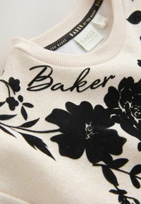 Cremefarbener Pullover mit schwarzem Blumenmuster und dem Text "Baker". Weicher Stoff, gerippter Kragen und subtiler Glanz im Material.