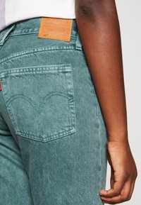 Primer plano de una persona que lleva puestos unos vaqueros Levi's 501 en color verde azulado, mostrando el bolsillo trasero, la etiqueta de la marca y su brazo derecho y mano relajados a un lado.