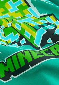 T-shirt grafica verde con un design a blocchi pixelati in giallo e blu, con la parola "MINECRAFT" in lettere nere in grassetto e verdi brillanti.
