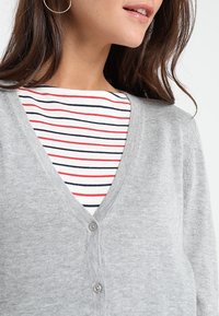 Cárdigan gris con un diseño de escote en V, que presenta un punto texturizado y dos botones. Debajo, se puede ver una camisa rayada en rojo, blanco y marino.