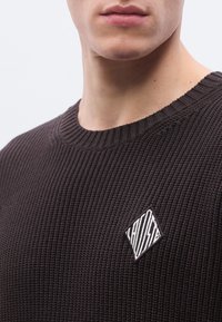Pull marron tricoté avec un design côtelé texturé et un petit patch losange brodé avec un motif rayé sur la poitrine.