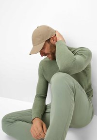 Beige Baseballkappe mit strukturiertem Design sitzt auf einer Person, die in einem grünen Strickpullover und passenden Hosen gekleidet ist, während sie vor einem weißen Hintergrund sitzt.