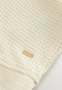 Beige texturerad tyg med ett upphöjt mönster, med en slät manschett och en guldig lapp märkt "DOWNTIME" fäst vid den.