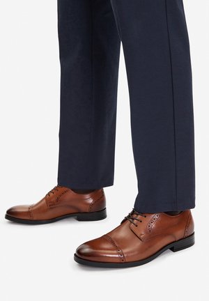 Pruunid nahast kleitkingad, millel on brogue detailid, paeltesüsteem ja läikiv viimistlus, koos tumesiniste pükstega.