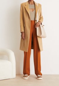 Cappotto camel, top in maglia beige con aperture, pantaloni arancioni a gamba larga e sneakers marroni. Include una borsa a spalla di colore chiaro.