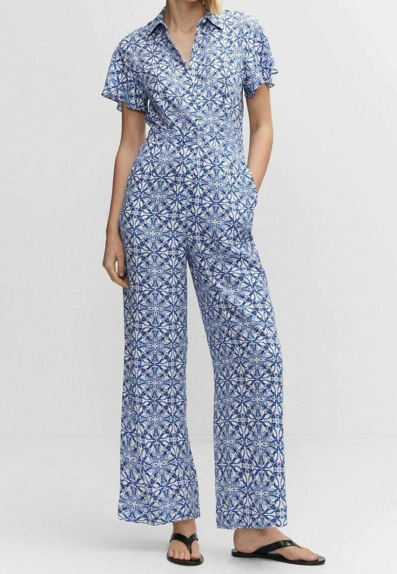 Femme portant une combinaison courte à manches courtes imprimée bleu et blanc avec des jambes larges et des sandales noires, debout avec une main dans la poche.