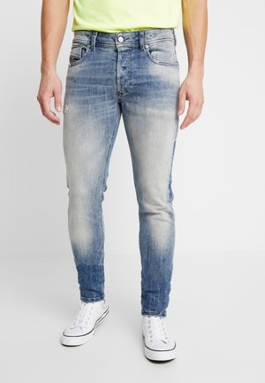 Vaqueros de denim azul desgastados con corte ajustado, que presentan áreas desgastadas y un lavado azul oscuro en la parte inferior de las piernas, combinados con zapatillas blancas.