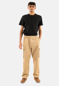 Beige cargo broek met elastische tailleband, twee zijzakken met rits en een relaxte pasvorm. Draagt met een zwart t-shirt met ronde hals en bruine schoenen.