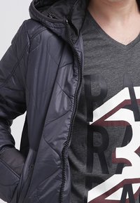 pikowana granatowa kurtka z kapturem, z zamkiem błyskawicznym z przodu i elastycznymi mankietami, noszona na szarym podkoszulku z dekoltem w serek z wyrazistym nadrukiem tekstowym.