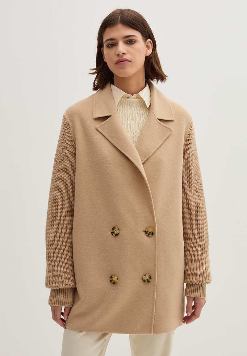 Beige Wolle Doppelreihermantel mit gestrickten Ärmeln. Verfügt über große runde Knöpfe und einen Reverskragen. Glatte Textur, oversized Schnitt.