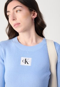 Svijetloplava rebrasta džemper s bijelim pravokutnim zakrpom s "CK" logotipom. Dodatak uključuje krem traku s torbe.