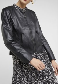 Černá kožená bomber bunda s kulatým límcem. Má zapínání na zip a dvě boční kapsy. Nosená přes vzorované šaty s bílými a černými tečkami.