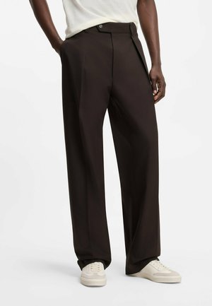 Mann trägt lockere schwarze maßgeschneiderte Hosen mit Seitentaschen und weißen Turnschuhen, steht mit einer Hand in der Tasche.