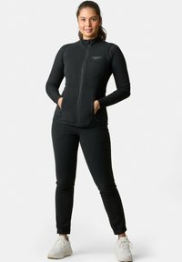 Sort zip-up atletisk jakke med ribbet tekstur og sidelommer, kombineret med matchende tætsiddende bukser og hvide sneakers.