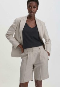 Beige linnen blazer over een zwart V-hals top, gecombineerd met bijpassende beige shorts met plooien en zijzakken. Soepele textuur.