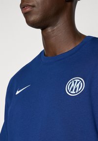 Kék pamut kereknyakú póló, fehér Nike logóval a mellkasán és kerek hímzett logóval, kék és fehér díszítéssel.
