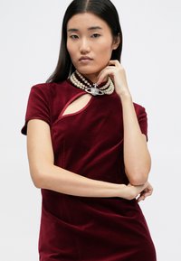 Bordowa sukienka z weluru z krótkimi rękawami, z wycięciem na klatce piersiowej. Uzupełniona wielowarstwowym naszyjnikiem z pereł.