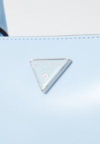 Borsa a tracolla blu chiaro realizzata in materiale sintetico liscio, con un logo a triangolo silver con la scritta "GUESS" e un design a punto interrogativo.