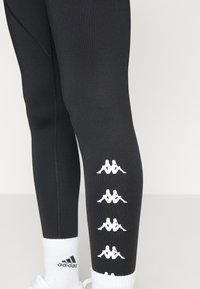 Mustat urheiluleggingsit, jotka on valmistettu stretch-kankaasta ja joissa on valkoiset Kappa-logo yksityiskohtia sekä sileä tekstuuri. Valkoiset sukat, joissa on Adidas-brändäys.