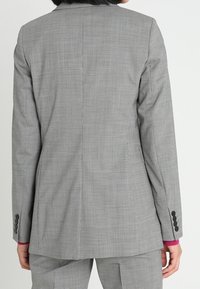 Šedá saka s dokonale padnoucím střihem, texturovaným vzorem, jedním rozparkem na zadní straně a černými knoflíky na rukávech.