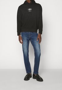 Svart hoodie med en logga framtill, kombinerad med blå denimjeans. Skor inkluderar svarta loafers med tjock sula. Enkel design.